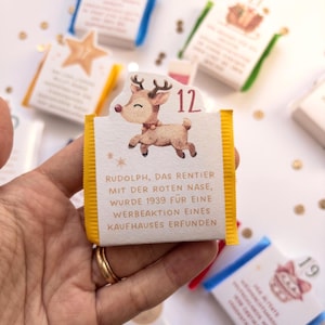 May include: A small rectangular chocolate bar wrapper with yellow side edges, featuring a cartoon reindeer illustration, the number 12, and German text stating Rudolph, das Rentier mit der roten Nase, wurde 1939 f&uuml;r eine Werbeaktion eines Kaufhauses erfunden.