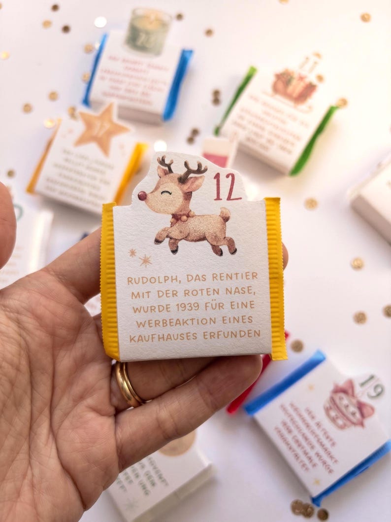 May include: A small rectangular chocolate bar wrapper with yellow side edges, featuring a cartoon reindeer illustration, the number 12, and German text stating Rudolph, das Rentier mit der roten Nase, wurde 1939 f&uuml;r eine Werbeaktion eines Kaufhauses erfunden.