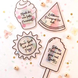 Op de afbeelding: Vier doorzichtige acrylvormen in de vorm van een cupcake, een watermeloenpartje, een zon en een ijsje, elk met een bankbiljet en confetti erin. De vormen hebben zwarte omlijningen en Duitse teksten waaronder Mach dir eine tolle Ferienzeit, Ein kleiner Feriengruß für dich, Sonne, Spaß und schöne Ferien, en Schöne Ferien und ganz viel Spaß.
