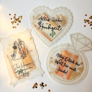 Op de afbeelding: Drie doorschijnende acrylhouders met gouden confetti en gevouwen biljetten van 50 euro. Eén is hartvormig met 'Alles Liebe zur Hochzeit', één is rechthoekig met een illustratie van een bruidspaar en één is ringvormig met 'Alles Glück der Welt'.