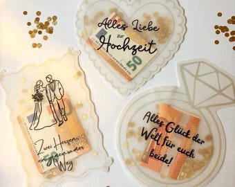 Könnte beinhalten: Drei durchscheinende Acrylhalter mit goldenem Konfetti und gefalteten 50-Euro-Scheinen. Einer ist herzförmig mit 'Alles Liebe zur Hochzeit', einer rechteckig mit Brautpaar-Illustration und einer ringförmig mit 'Alles Glück der Welt für euch beide!'.