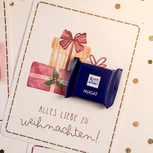 Op de afbeelding: Een kleine, vierkante, donkerblauwe Ritter Sport Nugat chocoladereep op een witte kaart met een aquarelillustratie van gestapelde cadeaus met strikken en de tekst Alles Liebe zu Weihnachten in bruine letters.