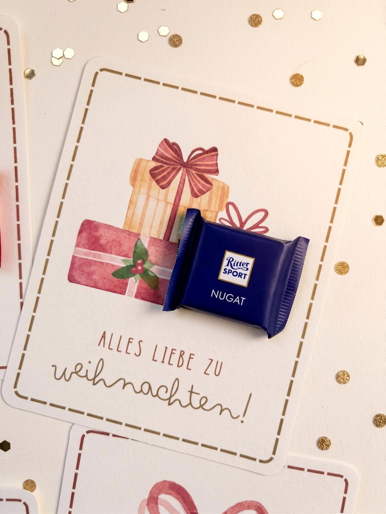 Op de afbeelding: Een kleine, vierkante, donkerblauwe Ritter Sport Nugat chocoladereep op een witte kaart met een aquarelillustratie van gestapelde cadeaus met strikken en de tekst Alles Liebe zu Weihnachten in bruine letters.