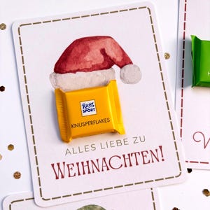 Op de afbeelding: Een rechthoekige witte kaart met een aquarelillustratie van een rode kerstmuts. Een geel Ritter Sport Knusperflakes chocoladevierkantje is op de kaart bevestigd. De tekst luidt ALLES LIEBE ZU WEIHNACHTEN in rode en grijze letters.
