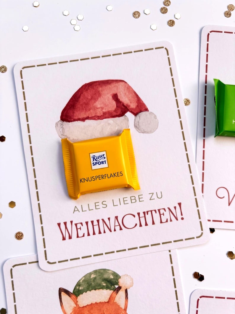 Op de afbeelding: Een rechthoekige witte kaart met een aquarelillustratie van een rode kerstmuts. Een geel Ritter Sport Knusperflakes chocoladevierkantje is op de kaart bevestigd. De tekst luidt ALLES LIEBE ZU WEIHNACHTEN in rode en grijze letters.