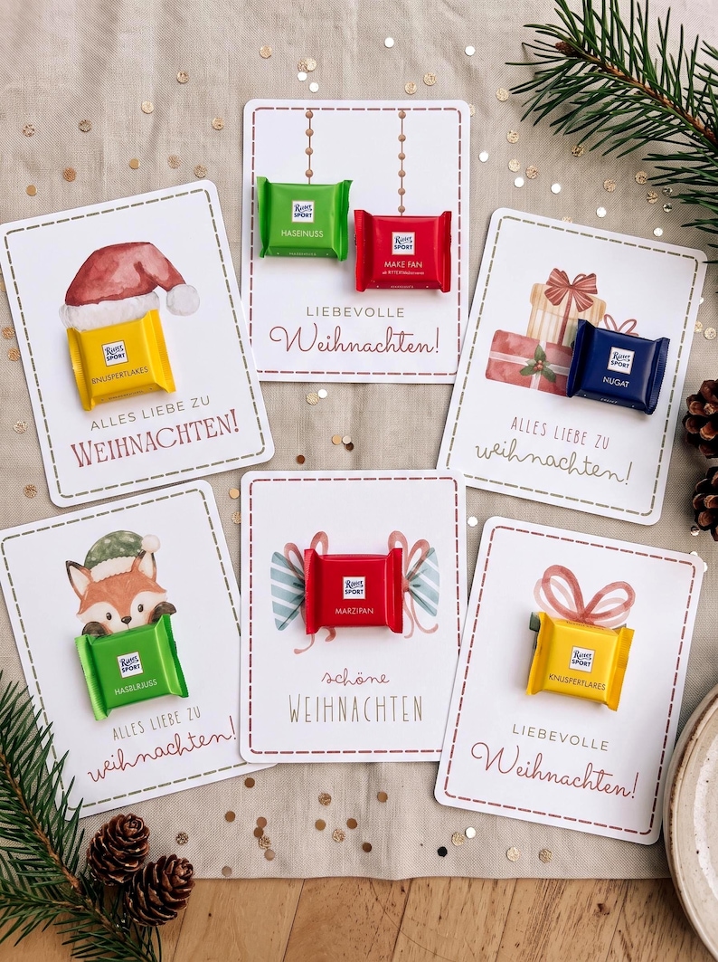 Op de afbeelding: Vijf rechthoekige witte kaarten met aquarelillustraties van kerstthema's, elk bevestigd met een klein Ritter Sport chocoladevierkantje. De tekst bevat 'ALLES LIEBE ZU WEIHNACHTEN!', 'LIEBEVOLLE Weihnachten!' en 'sch&ouml;ne WEIHNACHTEN'.
