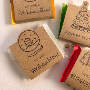 May include: Small rectangular chocolate bars wrapped in brown kraft paper with black line art illustrations of snowmen, Christmas trees, and snow globes. The wrappers feature German text including Sch&ouml;ne Weihnachten and Frohe Weihnachten.