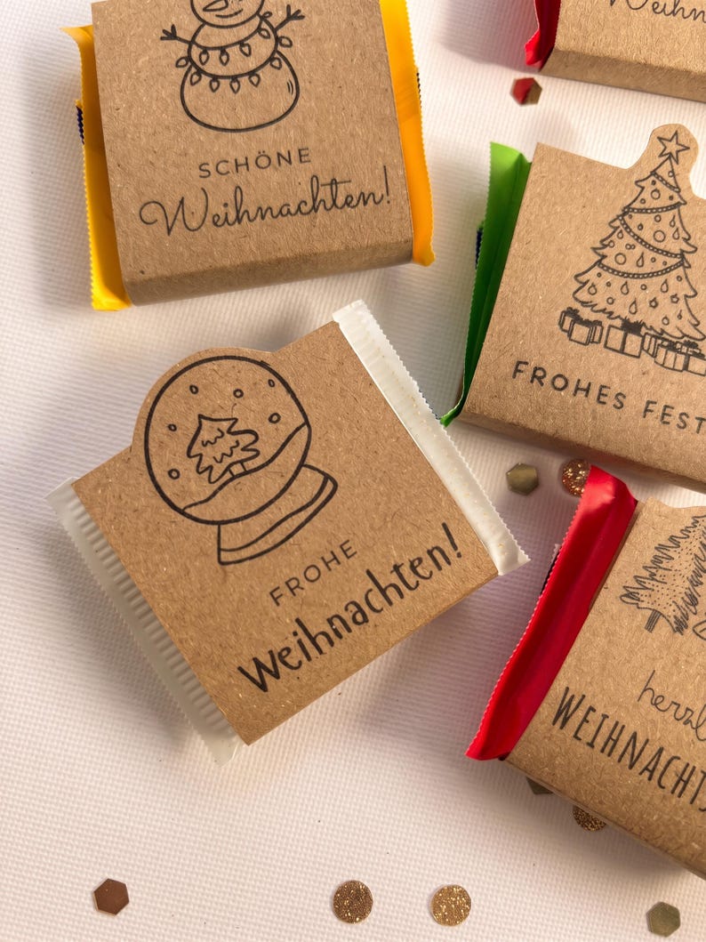 May include: Small rectangular chocolate bars wrapped in brown kraft paper with black line art illustrations of snowmen, Christmas trees, and snow globes. The wrappers feature German text including Sch&ouml;ne Weihnachten and Frohe Weihnachten.