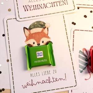 Op de afbeelding: Een witte rechthoekige kaart met een getekende vos met een groene muts, waarop een groene Ritter Sport Haselnuss chocoladereep is bevestigd. De kaart bevat de tekst ALLES LIEBE ZU WEIHNACHTEN! in donkerrode en zwarte letters.