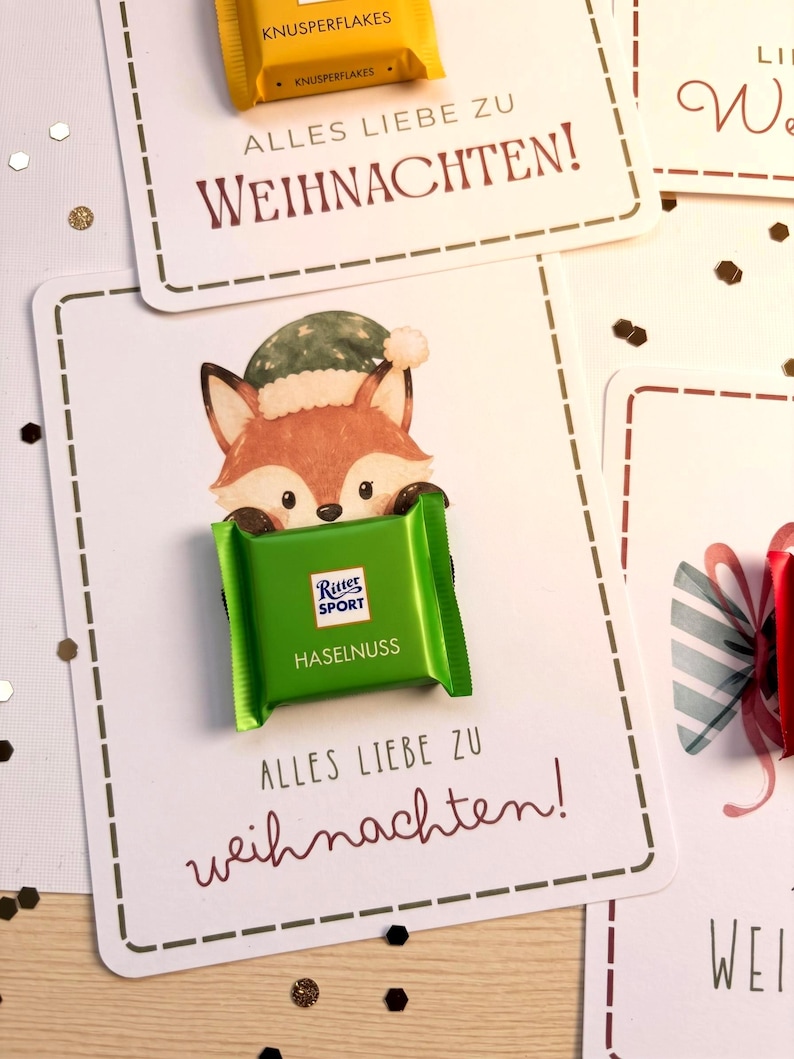 Op de afbeelding: Een witte rechthoekige kaart met een getekende vos met een groene muts, waarop een groene Ritter Sport Haselnuss chocoladereep is bevestigd. De kaart bevat de tekst ALLES LIEBE ZU WEIHNACHTEN! in donkerrode en zwarte letters.