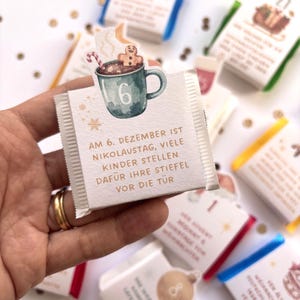 May include: A small square chocolate bar wrapper featuring a watercolor illustration of a teal mug with a gingerbread man and candy cane. The text reads: AM 6. DEZEMBER IST NIKOLAUSTAG, VIELE KINDER STELLEN DAF&Uuml;R IHRE STIEFEL VOR DIE T&Uuml;R.