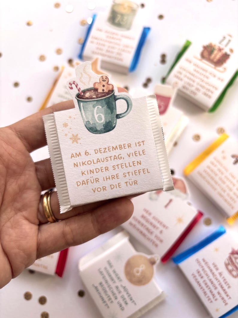 May include: A small square chocolate bar wrapper featuring a watercolor illustration of a teal mug with a gingerbread man and candy cane. The text reads: AM 6. DEZEMBER IST NIKOLAUSTAG, VIELE KINDER STELLEN DAF&Uuml;R IHRE STIEFEL VOR DIE T&Uuml;R.
