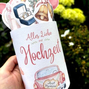 May include: A cylindrical white card featuring the German text Alles Liebe zur Hochzeit in pink script. The card is decorated with watercolor illustrations of a red car with a Just Married sign, gold wedding bells, rings in a blue box, and flowers.