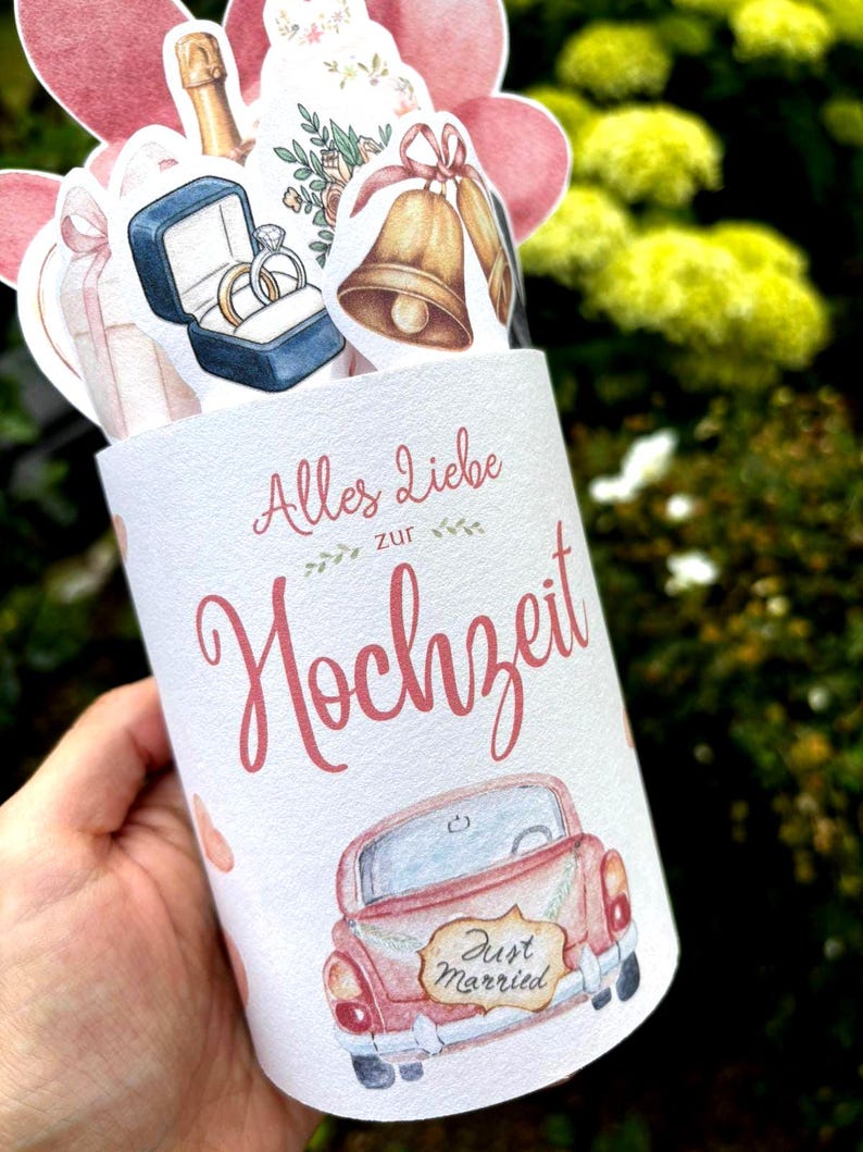 May include: A cylindrical white card featuring the German text Alles Liebe zur Hochzeit in pink script. The card is decorated with watercolor illustrations of a red car with a Just Married sign, gold wedding bells, rings in a blue box, and flowers.