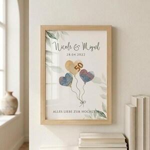 May include: Framed print with a light wood frame. The print features the names "Nicole & Miguel" above the date "28.04.2022". Three heart-shaped balloons are depicted, one with the number "50". The phrase "ALLES LIEBE ZUR HOCHZEITEN" is at the bottom.