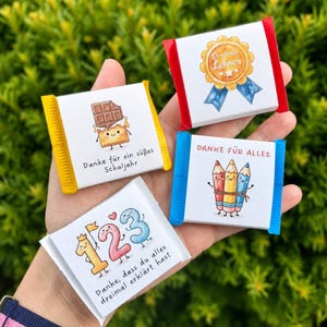 Op de afbeelding: Vier kleine rechthoekige chocoladerepen met witte wikkels voorzien van kleurrijke illustraties en Duitse tekst. De afbeeldingen tonen een chocoladereep, een gouden medaille, drie kleurpotloden en de cijfers 1, 2 en 3 met een kroontje.