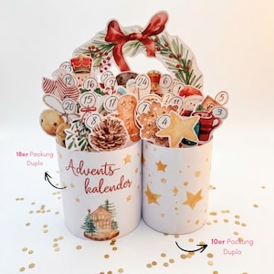 May include: A two-part cylindrical advent calendar featuring festive watercolor illustrations of a nutcracker, gingerbread man, pinecone, and star. The left cylinder displays the text Advents kalender, 18er Packung Duplo, and 10er Packung Duplo.