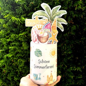 May include: A cylindrical white container featuring the text "Sch&ouml;ne Sommerferien!" and small summer-themed illustrations. A decorative topper displays a palm tree, a beach sign, a coconut drink, a starfish, and a slice of watermelon.