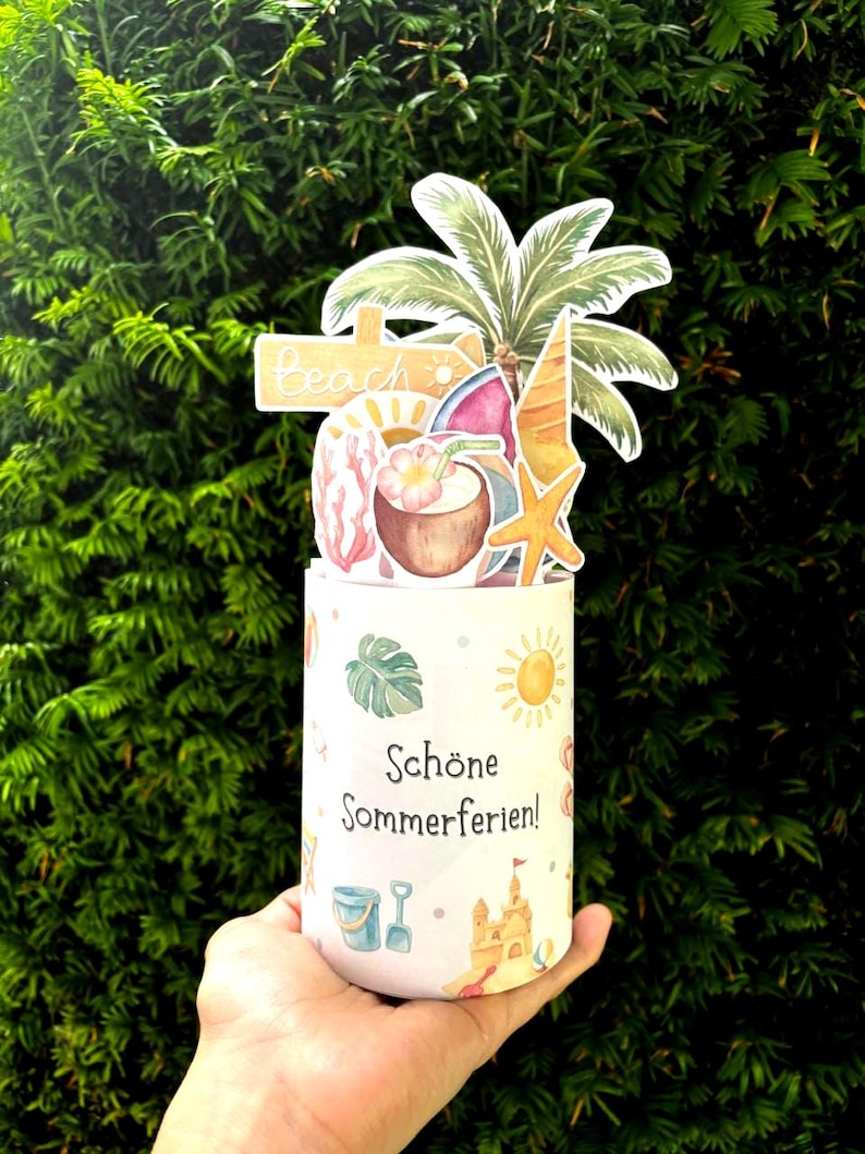 May include: A cylindrical white container featuring the text "Sch&ouml;ne Sommerferien!" and small summer-themed illustrations. A decorative topper displays a palm tree, a beach sign, a coconut drink, a starfish, and a slice of watermelon.