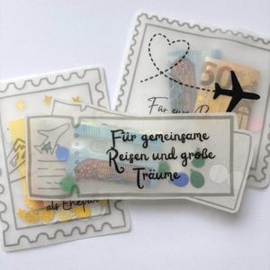 May include: Three translucent envelopes shaped like postage stamps and a ticket, containing paper currency and colorful confetti. The ticket features the black text "F&uuml;r gemeinsame Reisen und gro&szlig;e Tr&auml;ume" and a small airplane icon, while others show a heart and plane.