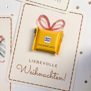 Op de afbeelding: Een kleine gele Ritter Sport Knusperflakes chocoladereep bevestigd op een rechthoekige witte kaart. De kaart bevat een aquarelillustratie van een rode strik en de tekst "LIEBEVOLLE Weihnachten!" in bruine letters.