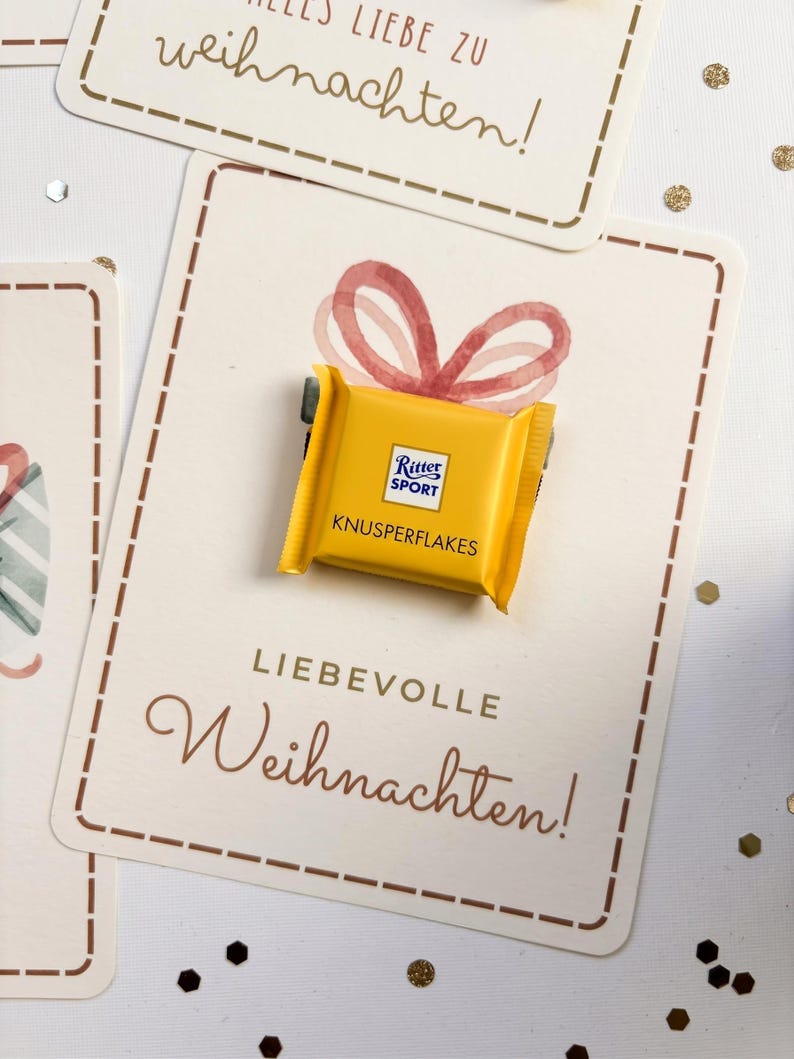 Op de afbeelding: Een kleine gele Ritter Sport Knusperflakes chocoladereep bevestigd op een rechthoekige witte kaart. De kaart bevat een aquarelillustratie van een rode strik en de tekst "LIEBEVOLLE Weihnachten!" in bruine letters.