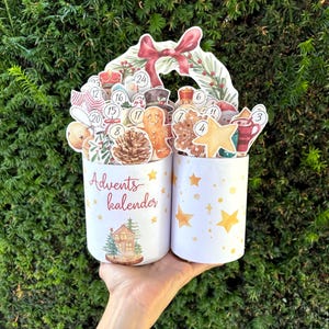 May include: Two white cylindrical containers featuring the text Advents kalender, decorated with yellow stars and a small house illustration. The containers are filled with various numbered Christmas-themed cutouts including a gingerbread man, pinecone, and star.