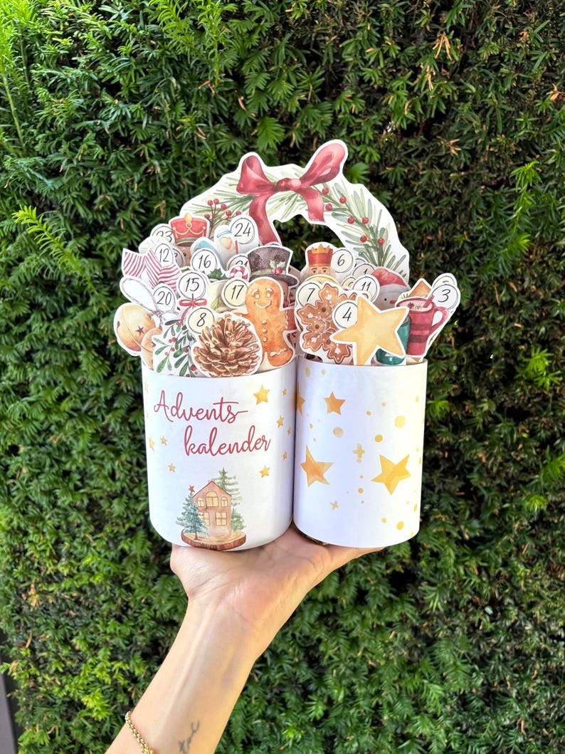 May include: Two white cylindrical containers featuring the text Advents kalender, decorated with yellow stars and a small house illustration. The containers are filled with various numbered Christmas-themed cutouts including a gingerbread man, pinecone, and star.