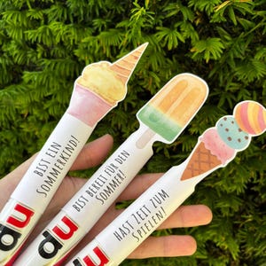 May include: Three white cylindrical tubes with red and black branding at the base, each topped with a different ice cream illustration. The text reads BIST EIN SOMMERKIND!, BIST BEREIT F&Uuml;R DEN SOMMER!, and HAST ZEIT ZUM SPIELEN! in black lettering.