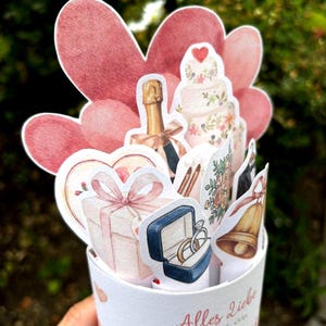 May include: A cylindrical paper card featuring a pop-up display of wedding-themed illustrations including a champagne bottle, a tiered cake, a gift box, a ring box with two rings, and a bell, set against pink heart cutouts with the text Alles Liebe zur Hochzeit.