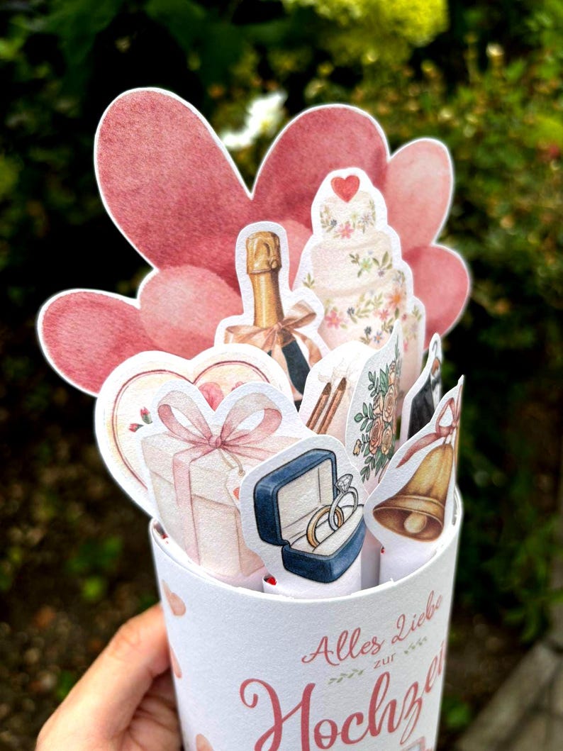 May include: A cylindrical paper card featuring a pop-up display of wedding-themed illustrations including a champagne bottle, a tiered cake, a gift box, a ring box with two rings, and a bell, set against pink heart cutouts with the text Alles Liebe zur Hochzeit.