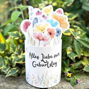 Puede incluir: Un recipiente cilíndrico blanco con el texto alemán "Alles Liebe zum Geburtstag" y un diseño floral. Contiene numerosos recortes de papel de coloridas flores de acuarela. El fondo presenta follaje verde.