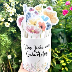 Puede incluir: Un recipiente cil&iacute;ndrico blanco con el texto negro Alles Liebe zum Geburtstag y un estampado floral en la base. El recipiente est&aacute; lleno de varias ilustraciones de flores troqueladas estilo acuarela en tonos rosa, amarillo y azul.