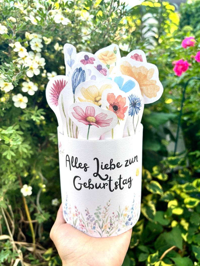 Puede incluir: Un recipiente cil&iacute;ndrico blanco con el texto negro Alles Liebe zum Geburtstag y un estampado floral en la base. El recipiente est&aacute; lleno de varias ilustraciones de flores troqueladas estilo acuarela en tonos rosa, amarillo y azul.