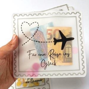 May include: A square, translucent envelope with a scalloped border, featuring a black airplane silhouette, a dashed heart flight path, and the German text "F&uuml;r eure Reise ins Gl&uuml;ck". Inside, a fifty euro banknote and pink circular confetti are visible.