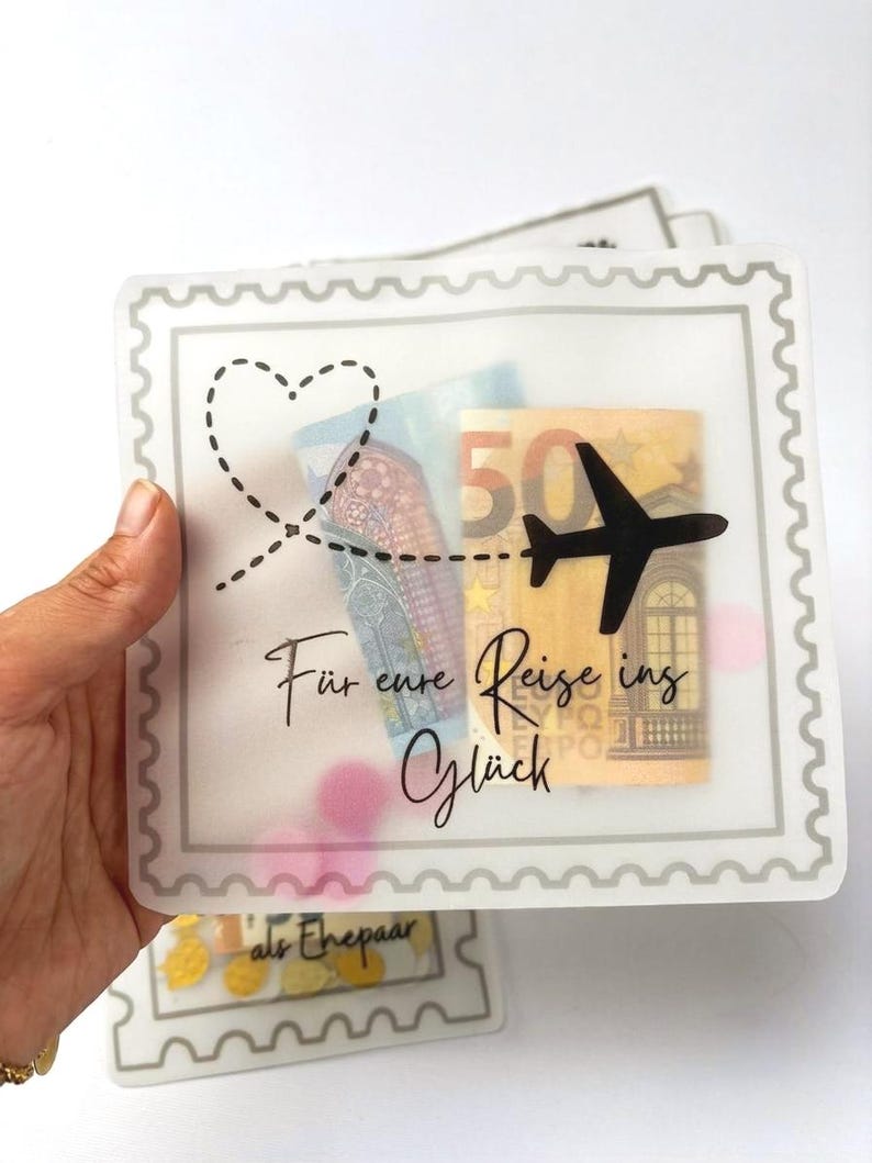 May include: A square, translucent envelope with a scalloped border, featuring a black airplane silhouette, a dashed heart flight path, and the German text "F&uuml;r eure Reise ins Gl&uuml;ck". Inside, a fifty euro banknote and pink circular confetti are visible.