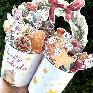 May include: Two white cylindrical containers featuring gold star patterns and the text Advents kalender. They are filled with various Christmas-themed cutouts including a gingerbread man, pinecone, snowflake, star, and numbered circles from 1 to 24.