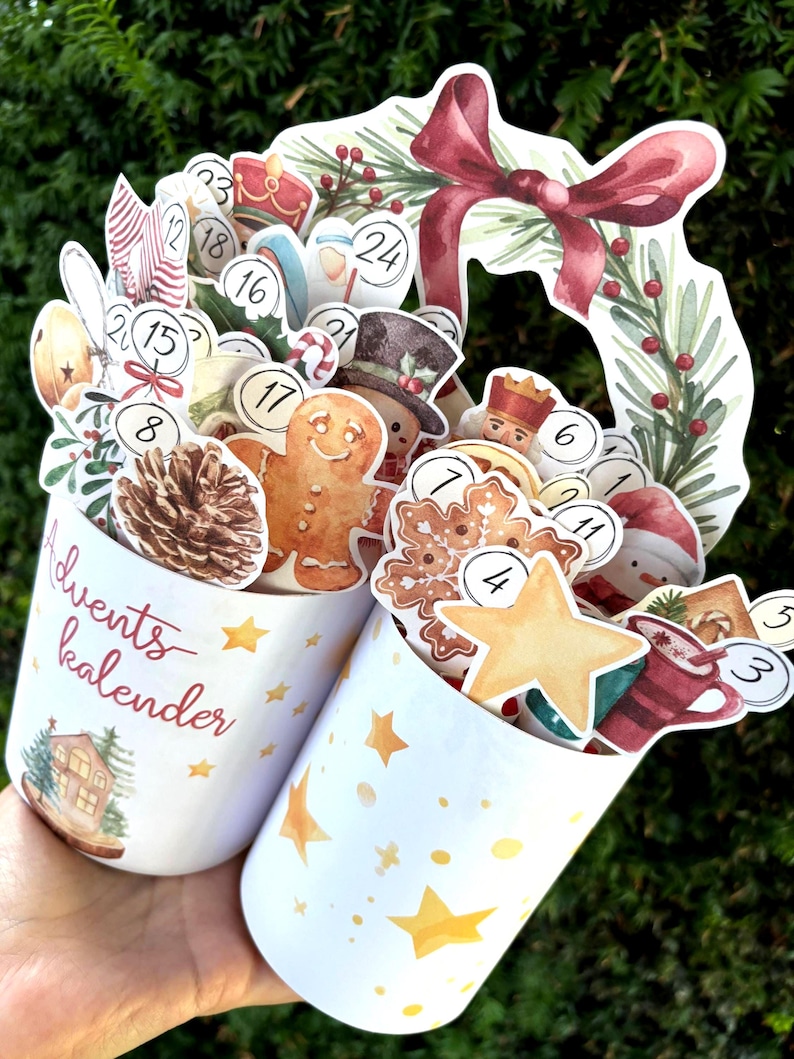 May include: Two white cylindrical containers featuring gold star patterns and the text Advents kalender. They are filled with various Christmas-themed cutouts including a gingerbread man, pinecone, snowflake, star, and numbered circles from 1 to 24.