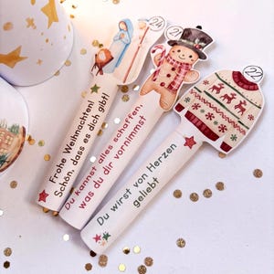 May include: Three white cylindrical items with flat tops featuring Christmas illustrations and German text. One shows a nativity scene with the number 24, another a gingerbread man with 21, and the third a patterned sweater with 22. Each has printed text.