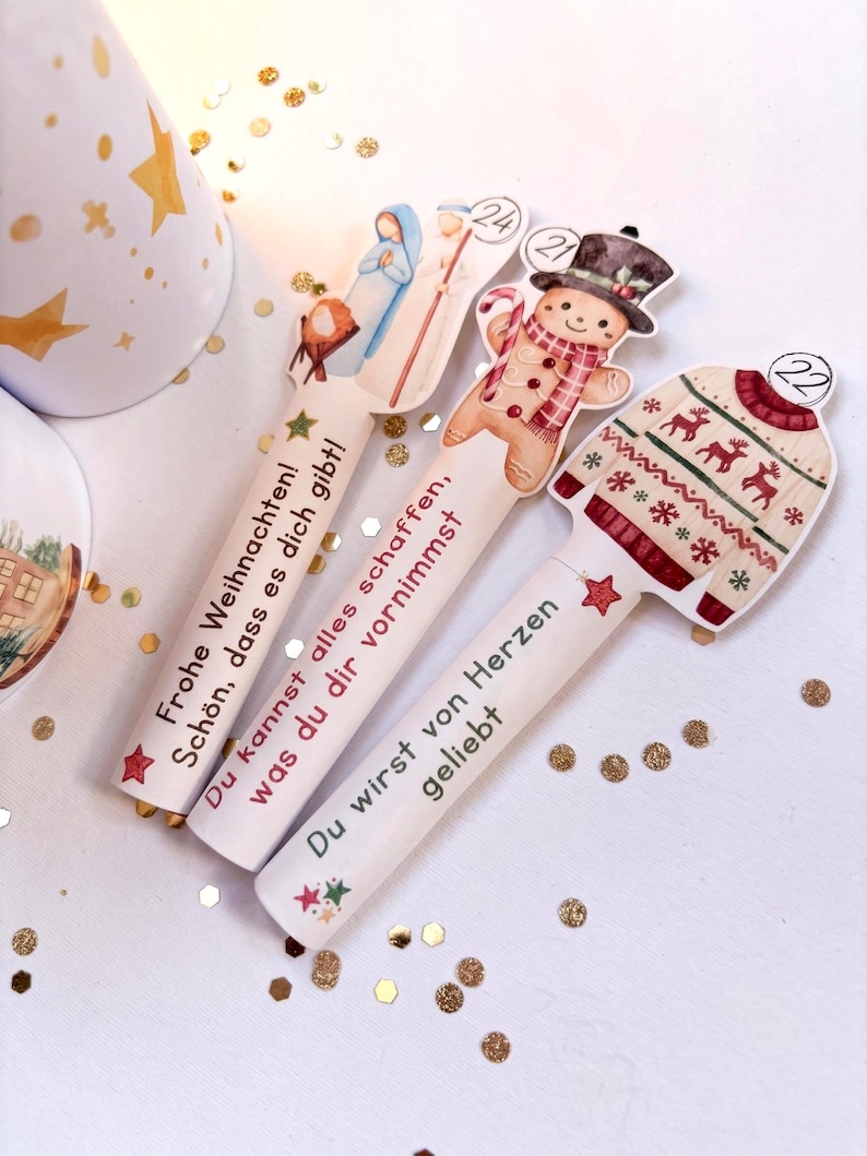 May include: Three white cylindrical items with flat tops featuring Christmas illustrations and German text. One shows a nativity scene with the number 24, another a gingerbread man with 21, and the third a patterned sweater with 22. Each has printed text.