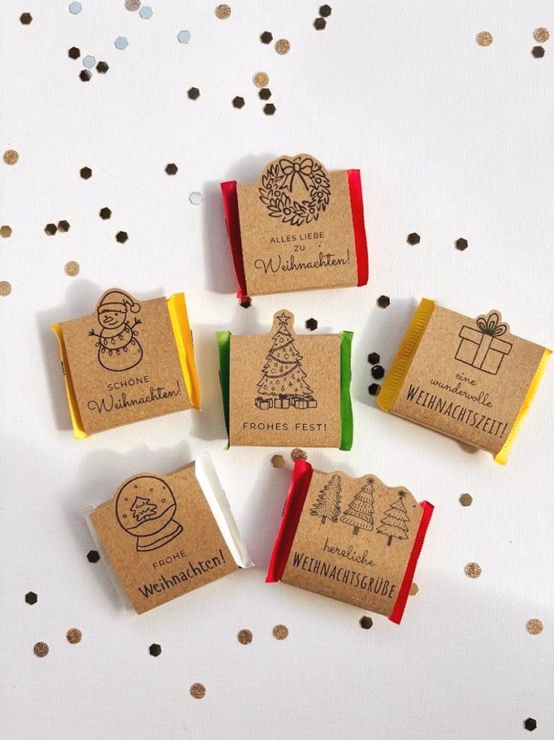 May include: Six small rectangular chocolate bars wrapped in brown kraft paper with festive black ink illustrations and German holiday text. Each wrapper features unique designs like a snowman, Christmas tree, wreath, gift, snow globe, and forest.