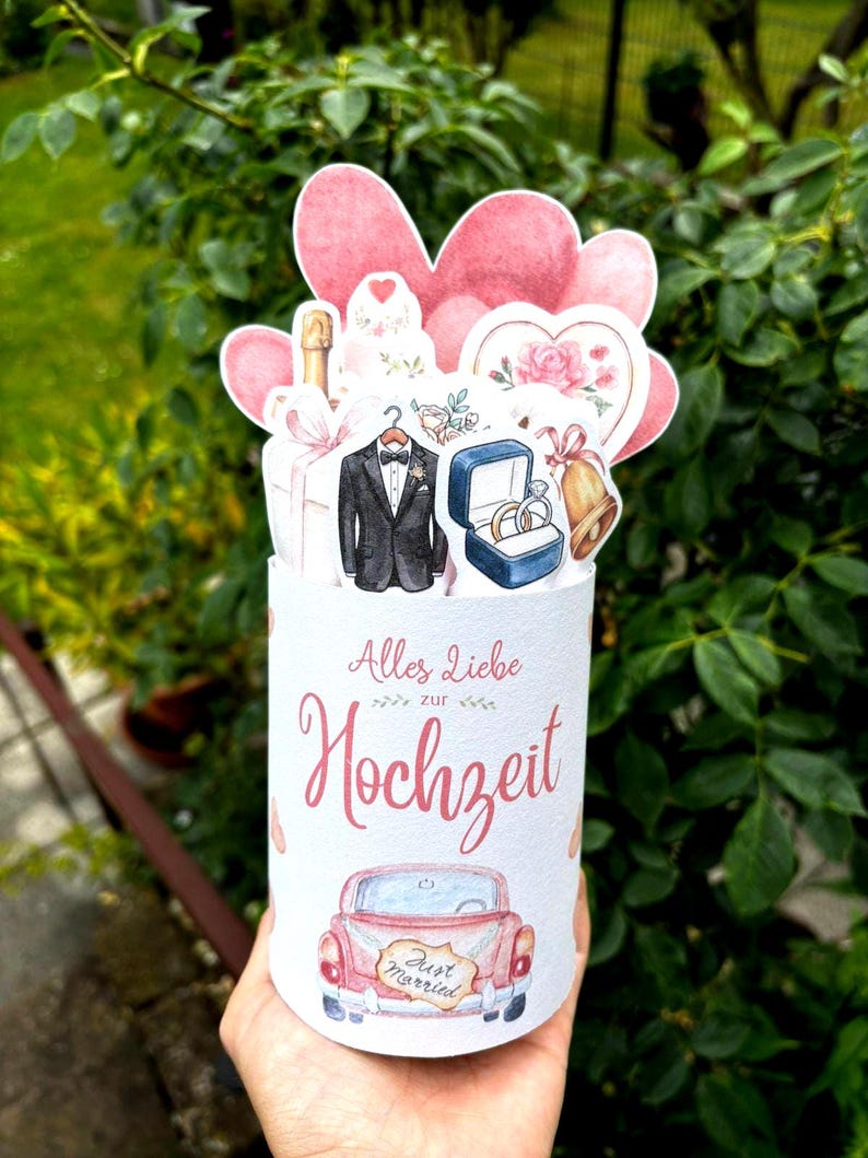 May include: A cylindrical wedding greeting card featuring the German text Alles Liebe zur Hochzeit. The design includes watercolor illustrations of a tuxedo, a ring box with two rings, a bell, a champagne bottle, and a pink car with a Just Married sign.