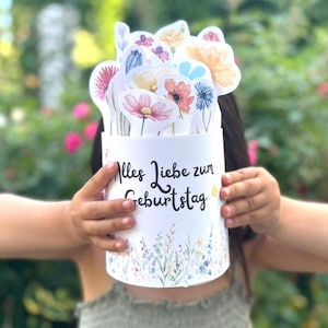 Puede incluir: Un recipiente cil&iacute;ndrico blanco con el texto en alem&aacute;n Alles Liebe zum Geburtstag en letra cursiva negra, decorado con un borde floral de acuarela en la base y lleno de varias ilustraciones de flores recortadas en acuarela.
