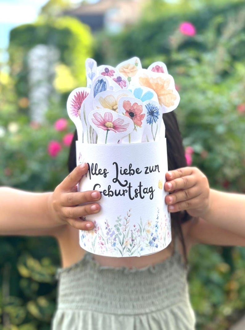 Puede incluir: Un recipiente cil&iacute;ndrico blanco con el texto en alem&aacute;n Alles Liebe zum Geburtstag en letra cursiva negra, decorado con un borde floral de acuarela en la base y lleno de varias ilustraciones de flores recortadas en acuarela.