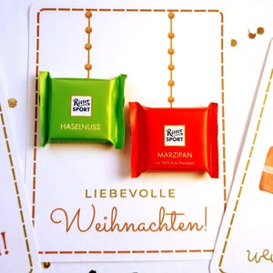 Op de afbeelding: Twee vierkante Ritter Sport chocoladerepen in groene en rode wikkels. De groene wikkel is gelabeld Haselnuss en de rode wikkel is gelabeld Marzipan met 100% Edel-Marzipan. Ze liggen op een witte kaart met de tekst Liebevolle Weihnachten.
