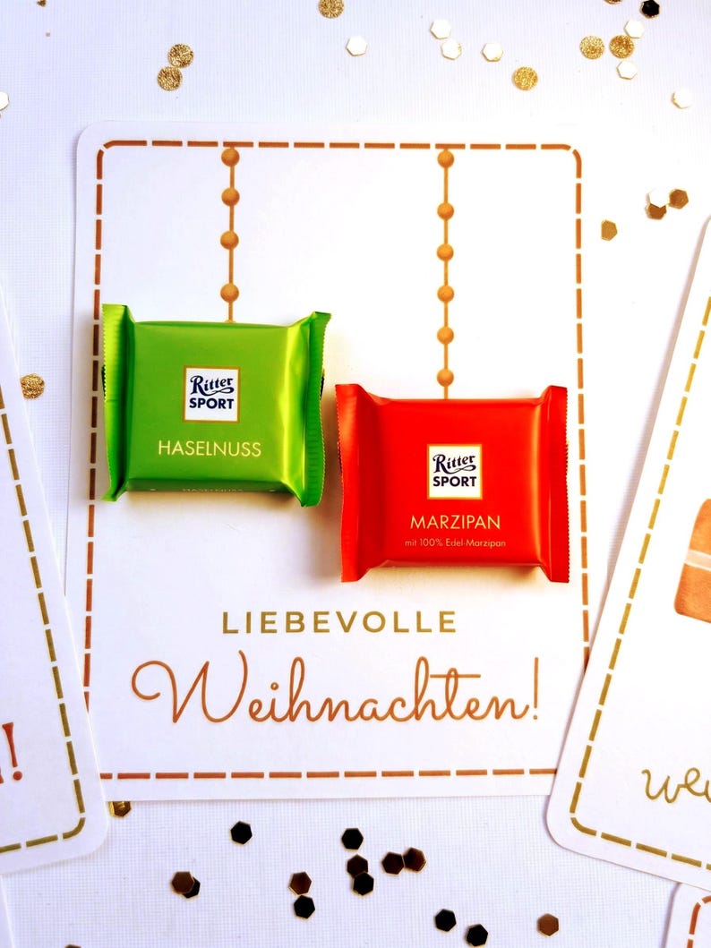 Op de afbeelding: Twee vierkante Ritter Sport chocoladerepen in groene en rode wikkels. De groene wikkel is gelabeld Haselnuss en de rode wikkel is gelabeld Marzipan met 100% Edel-Marzipan. Ze liggen op een witte kaart met de tekst Liebevolle Weihnachten.