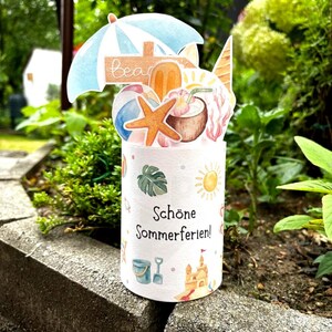 May include: A cylindrical paper card featuring the German text Sch&ouml;ne Sommerferien. The top displays a pop-up arrangement of a blue beach umbrella, a starfish, a coconut drink, a popsicle, and a beach ball. The base shows small summer icons.