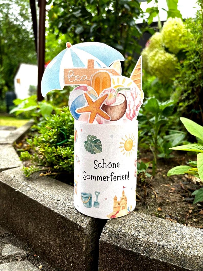 May include: A cylindrical paper card featuring the German text Sch&ouml;ne Sommerferien. The top displays a pop-up arrangement of a blue beach umbrella, a starfish, a coconut drink, a popsicle, and a beach ball. The base shows small summer icons.