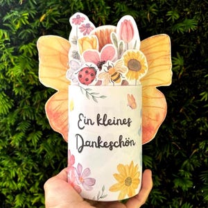 May include: A cylindrical container featuring large orange butterfly wings on the sides and a floral arrangement on top. The front displays the German text "Ein kleines Dankeschön" surrounded by watercolor illustrations of flowers, a ladybug, and a bee.