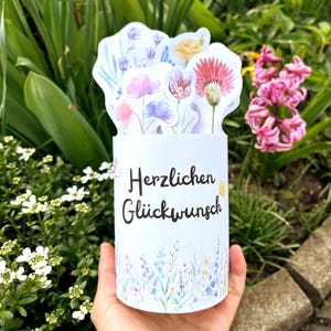 Könnte beinhalten: Eine zylindrische weiße Grußkarte mit dem schwarzen Text Herzlichen Glückwunsch auf der Vorderseite. Die Karte zeigt ein buntes Aquarell-Blumenmuster am unteren Rand und enthält mehrere ausgestanzte Blumenillustrationen am oberen Ende.
